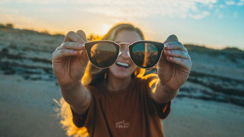 Women holding a pair of wooden sunglasses up on a beach during a sunset | Vilo Eyewear Australia